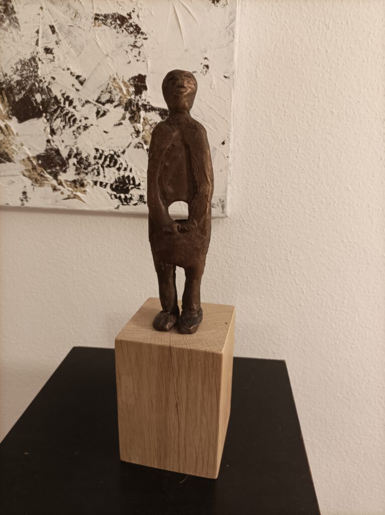 Skulptur Gottfried Sandböck: In der Mitte SEIN. Eine abstrakt dargestellte Person mit Loch im Bauch, die Arme sind vor dem Loch gefaltet. Die Figur steht auf einem Holzsockel. Kunst