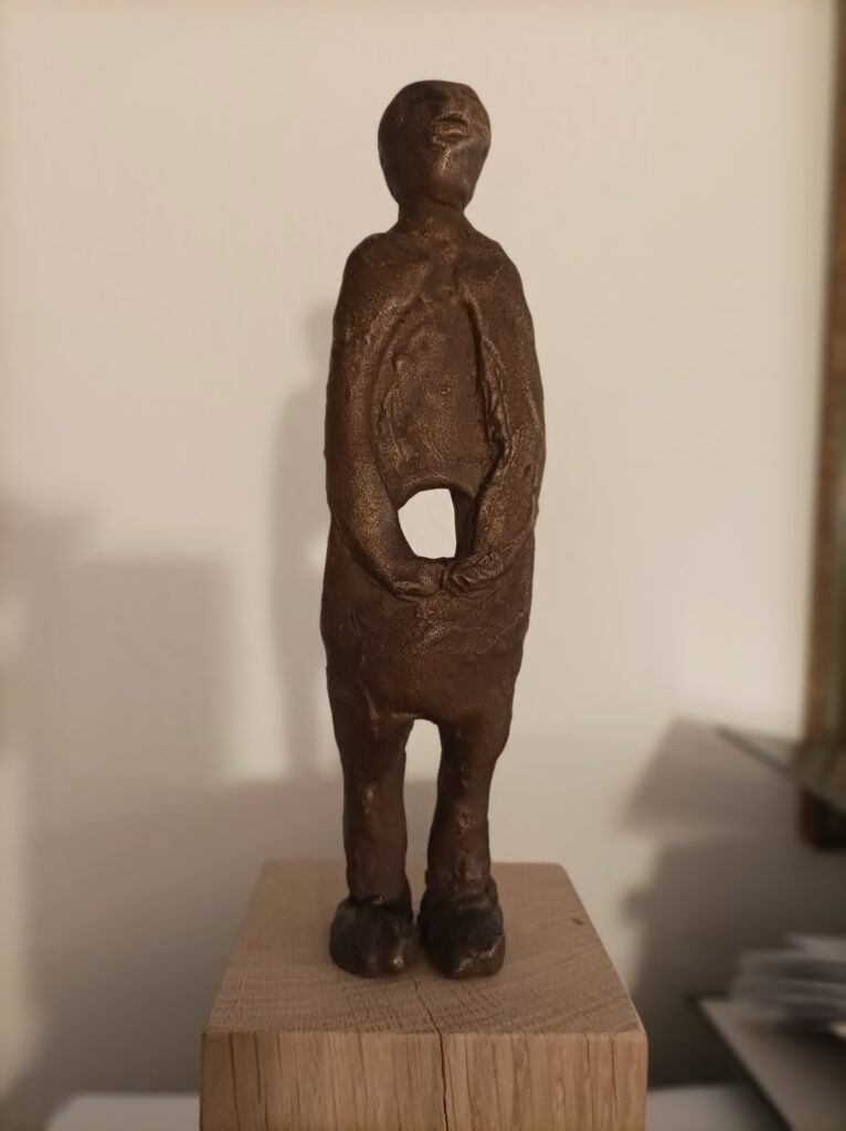 Skulptur Gottfried Sandböck: In der Mitte SEIN. Eine abstrakt dargestellte Person mit Loch im Bauch, die Arme sind vor dem Loch gefaltet. Die Figur steht auf einem Holzsockel. Kunst
