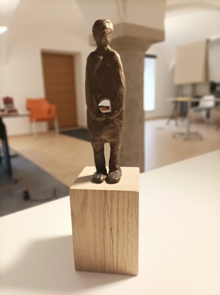 Skulptur Gottfried Sandböck: In der Mitte SEIN. Eine abstrakt dargestellte Person mit Loch im Bauch, die Arme sind vor dem Loch gefaltet. Die Figur steht auf einem Holzsockel.