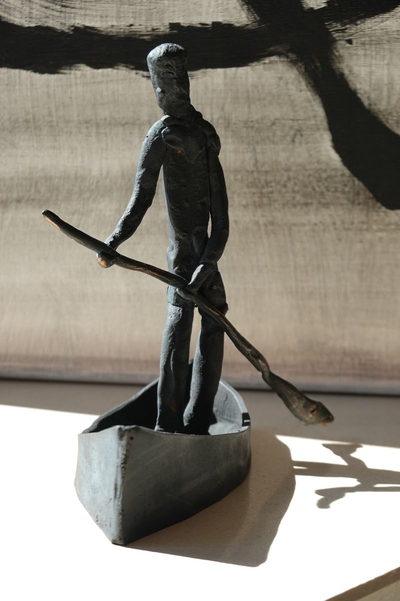 Schwarz patinierte Bronze-Skulptur einer stehenden Figur in einem Boot mit Ruder, im Hintergrund hängt ein abstraktes Gemälde.