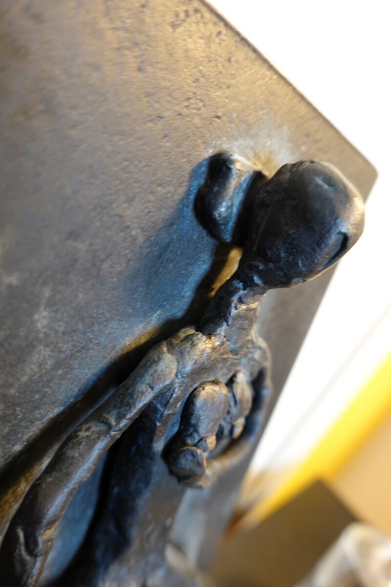 Schwarz patinierte Bronze-Skulptur einer stehenden Figur in einem Boot mit Ruder, im Hintergrund hängt ein abstraktes Gemälde.