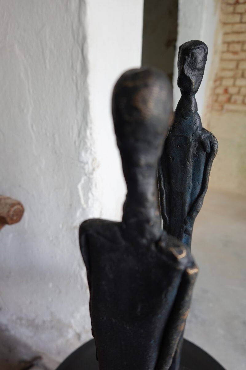 Schwarz patinierte Bronze-Skulptur einer stehenden Figur in einem Boot mit Ruder, im Hintergrund hängt ein abstraktes Gemälde.